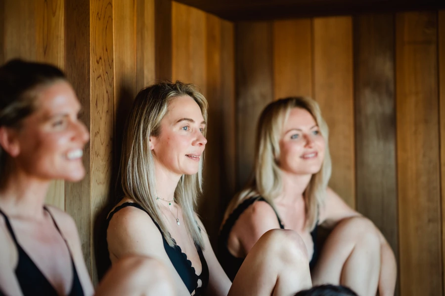 Three women sitting and relaxing inside a wooden sauna cabin.