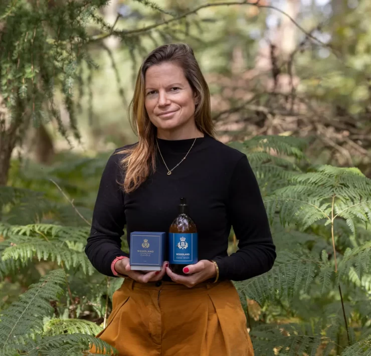 Laura Thomas, founder of Laura Thomas Co, holding the Balmoral Estate Woodland candle and hand wash surrounded by woodland greenery.