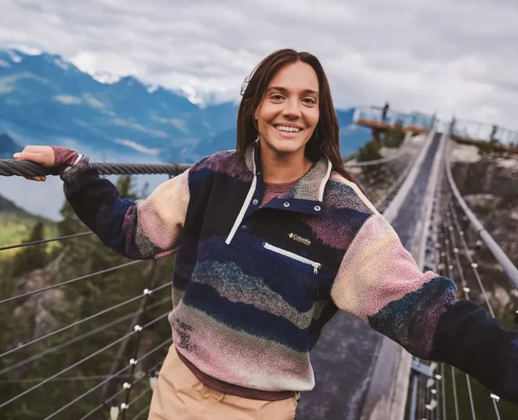 Woman wearing a colourful Columbia fleece outdoors on a mountain bridge, showcasing the brand’s performance outerwear collection.