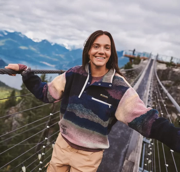 Woman wearing a colourful Columbia fleece outdoors on a mountain bridge, showcasing the brand’s performance outerwear collection.