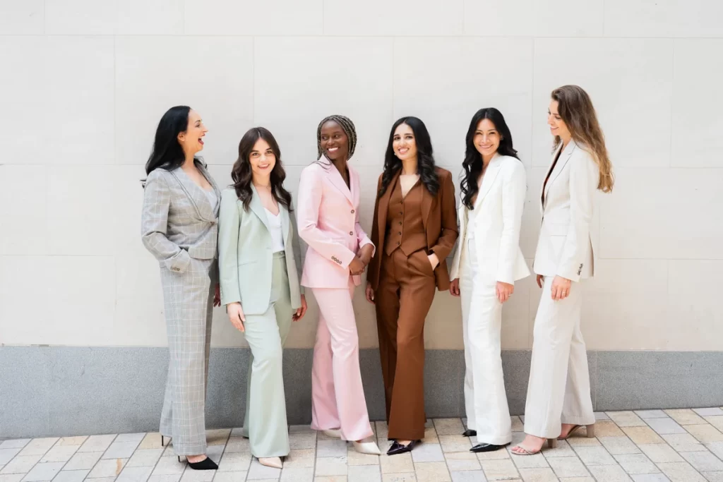 Six women wearing Gormley & Gamble tailored suits in pastel and neutral tones standing together outside on a stone walkway