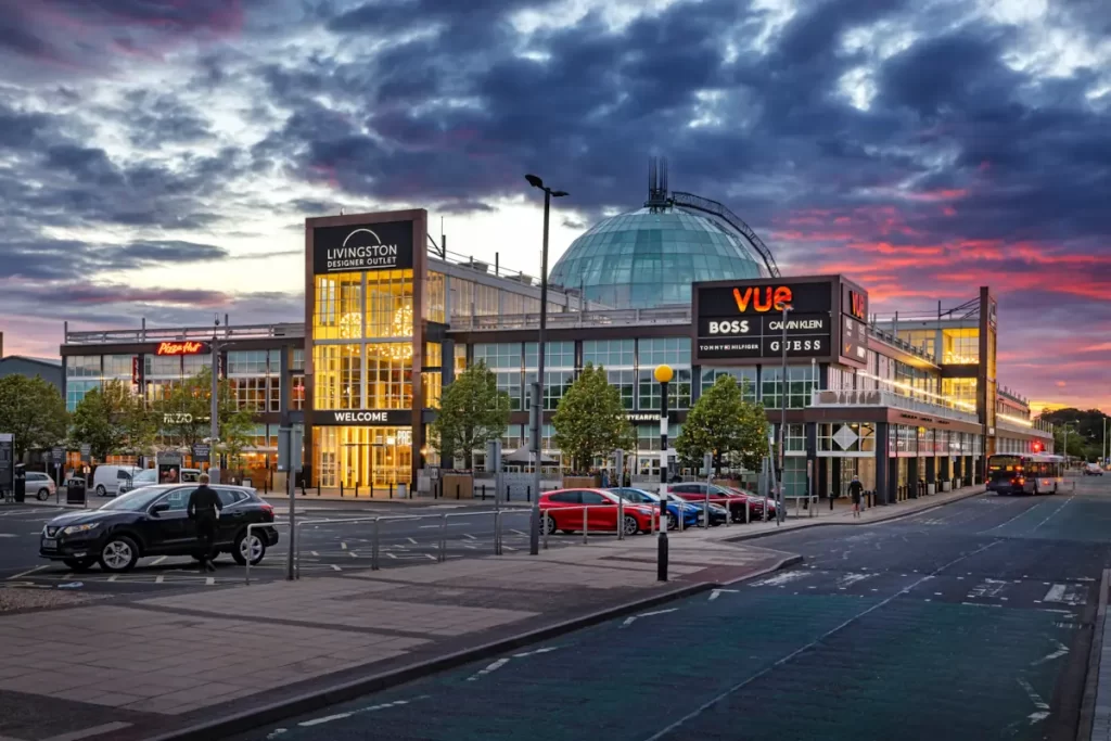 Exterior view of Livingston Designer Outlet at sunset with Vue cinema and fashion brand signage visible.