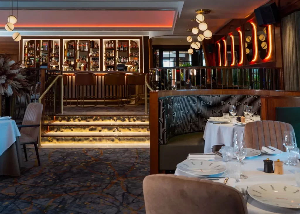 Art deco style restaurant and bar interior at Trocadero’s, Crossbasket Castle in Scotland, featuring patterned banquette seating, round tables set for dining, and a glowing backlit bar.