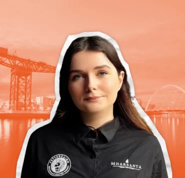 Portrait of Mae MacInnes, general manager of Mharsanta in Glasgow, wearing a branded shirt with an orange-toned background featuring the River Clyde, Finnieston Crane and Squinty Bridge.
