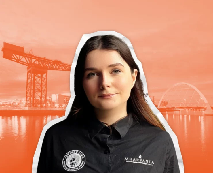 Portrait of Mae MacInnes, general manager of Mharsanta in Glasgow, wearing a branded shirt with an orange-toned background featuring the River Clyde, Finnieston Crane and Squinty Bridge.