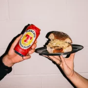 Two hands holding a Tennent’s Bavarian Pilsner can and a Square Wurst roll from Auld Hag against a white wall.