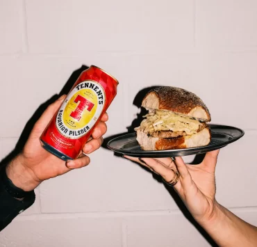 Two hands holding a Tennent’s Bavarian Pilsner can and a Square Wurst roll from Auld Hag against a white wall.