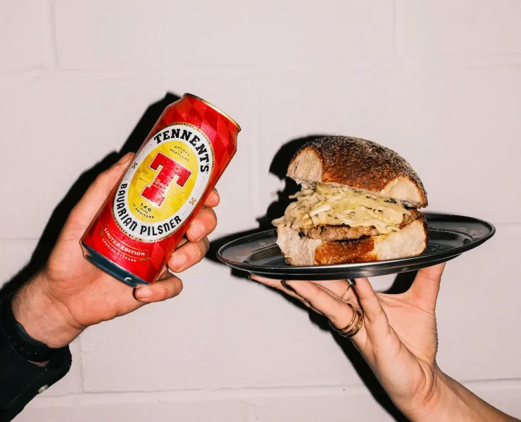 Two hands holding a Tennent’s Bavarian Pilsner can and a Square Wurst roll from Auld Hag against a white wall.