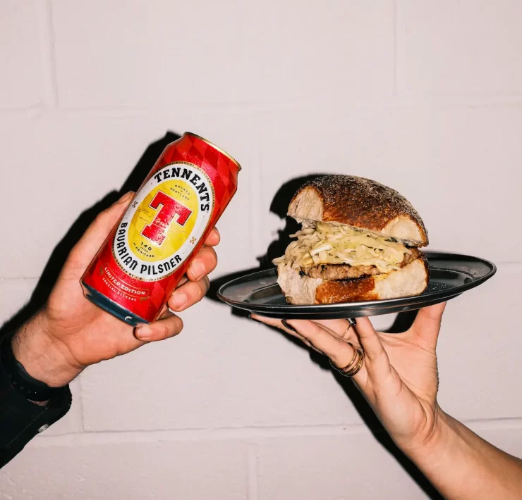 Two hands holding a Tennent’s Bavarian Pilsner can and a Square Wurst roll from Auld Hag against a white wall.