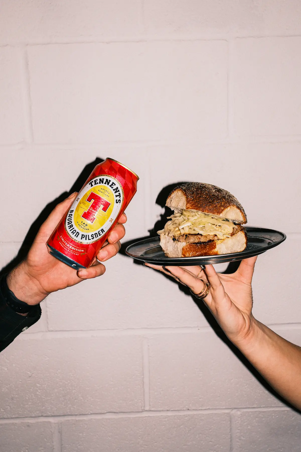Two hands holding a Tennent’s Bavarian Pilsner can and a Square Wurst roll from Auld Hag against a white wall.