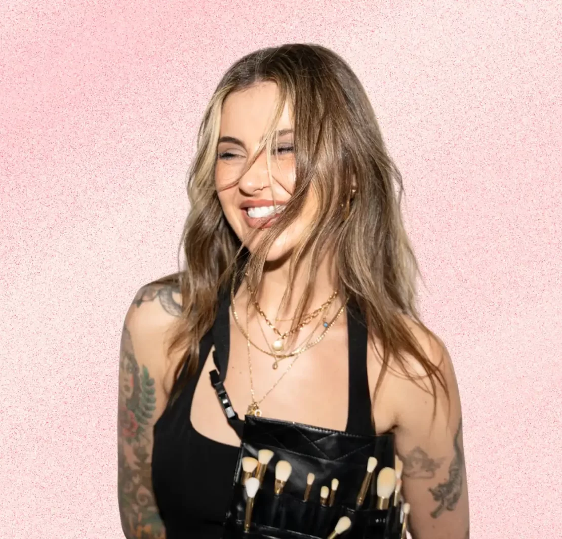 Jamie Genevieve smiling against a soft pink textured background, wearing layered gold jewellery and a makeup artist brush belt.