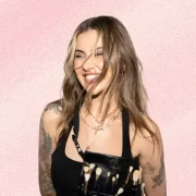 Jamie Genevieve smiling against a soft pink textured background, wearing layered gold jewellery and a makeup artist brush belt.