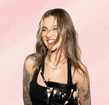 Jamie Genevieve smiling against a soft pink textured background, wearing layered gold jewellery and a makeup artist brush belt.