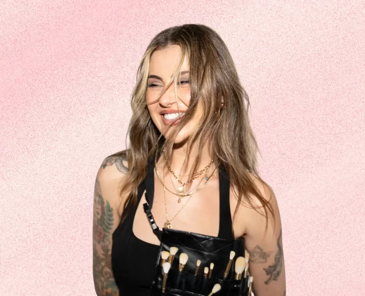 Jamie Genevieve smiling against a soft pink textured background, wearing layered gold jewellery and a makeup artist brush belt.