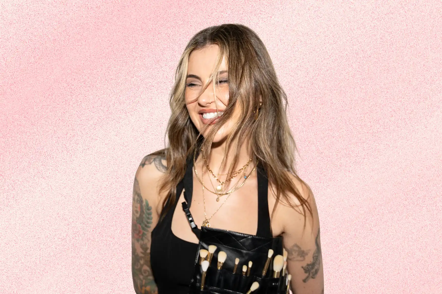 Jamie Genevieve smiling against a soft pink textured background, wearing layered gold jewellery and a makeup artist brush belt.