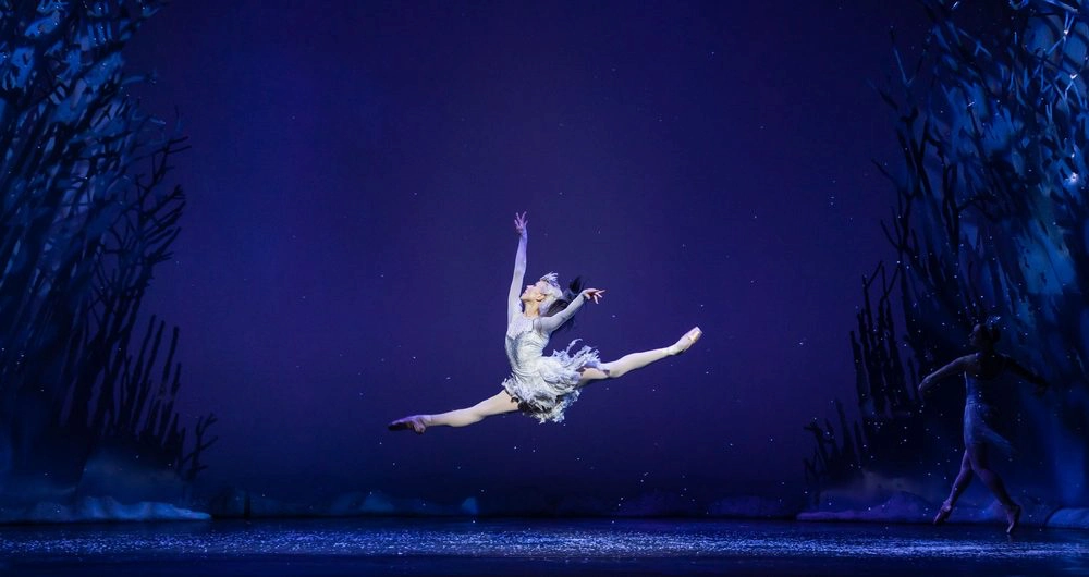 Ballet dancer Constance Devernay leaps across the stage as the Snow Queen, surrounded by an icy, winter-themed set.