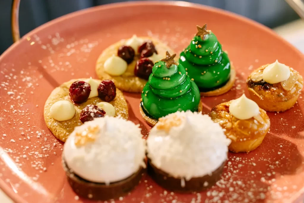 A close-up of festive afternoon tea desserts at Champagne Central Glasgow, featuring cherry-topped biscuits, green Christmas tree-shaped pastries, snowball-style cakes and mini mince bite tarts on a pink plate.