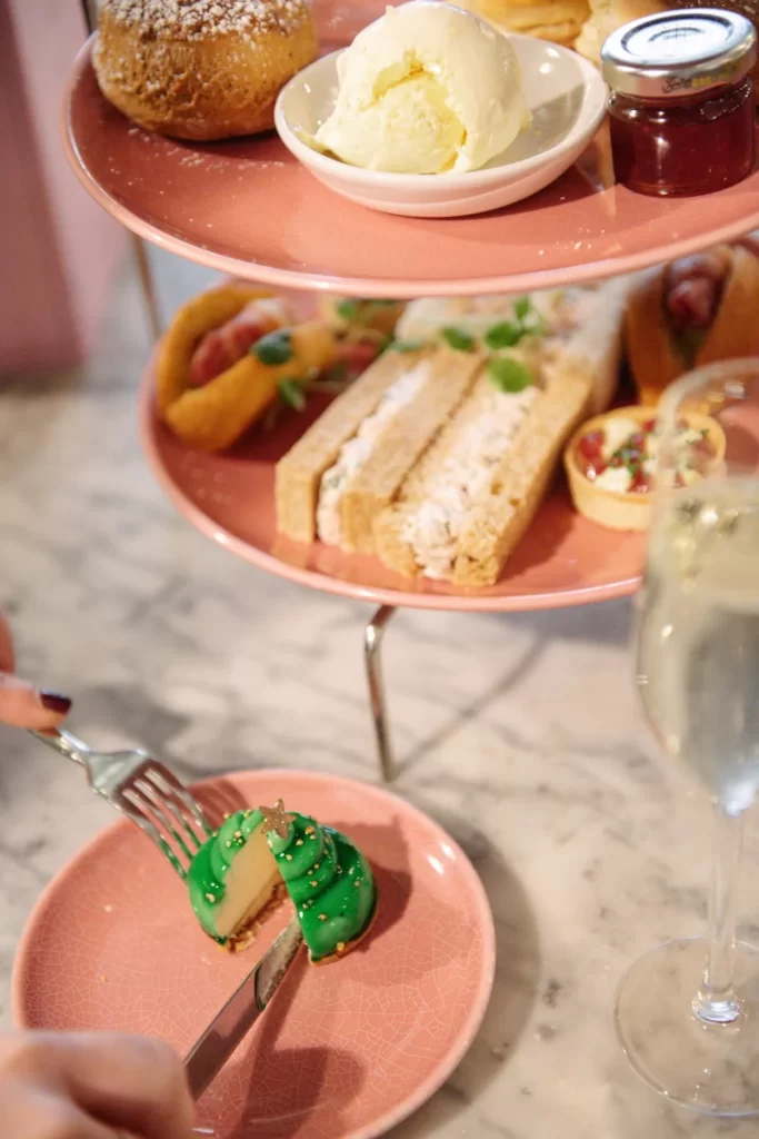 This image shows a detailed view of the festive afternoon tea at Champagne Central in voco Grand Central Glasgow. A pink tiered stand holds scones, clotted cream, jam, savoury bites and delicate finger sandwiches, while a guest cuts into a Christmas tree-shaped dessert on a matching pink plate. The soft marble table and a glass of fizz add to the seasonal, elegant feel. Ideal for articles highlighting Glasgow’s festive dining experiences.