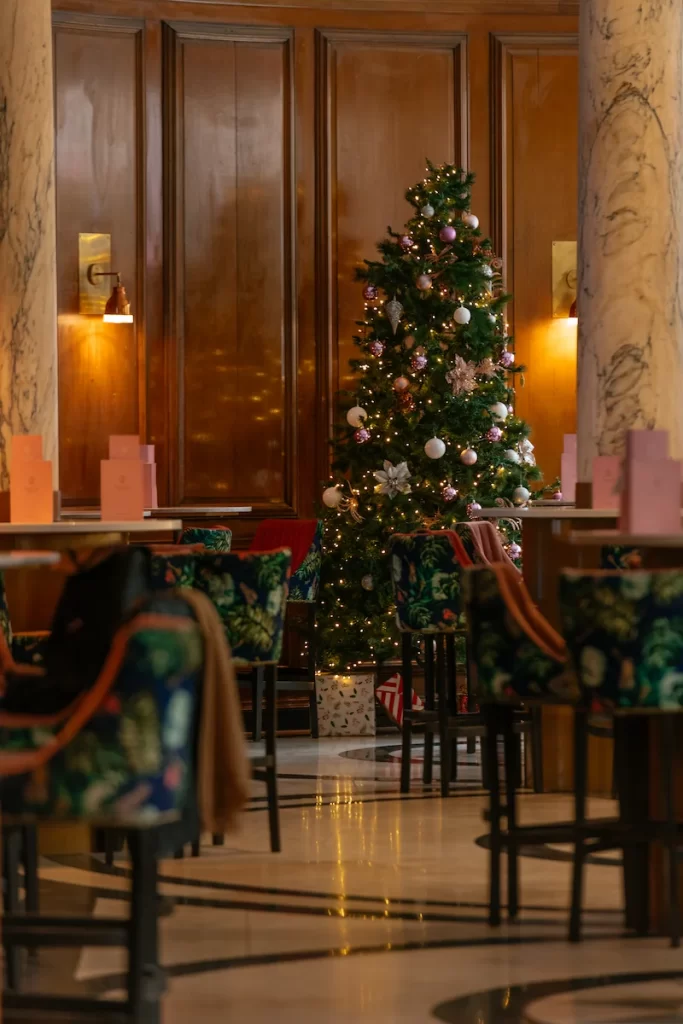 A Christmas tree decorated with lights and ornaments inside Champagne Central at voco Grand Central Glasgow, surrounded by patterned velvet chairs and warm wooden panelling.