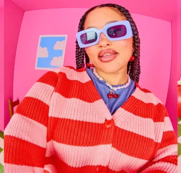 Models wearing colourful Lucy & Yak cardigans, knitwear and graphic outfits in a bright pink studio.