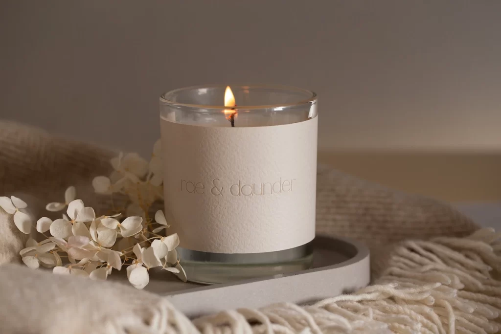 A lit rae & daunder candle placed on a tray with dried flowers and soft beige blankets.