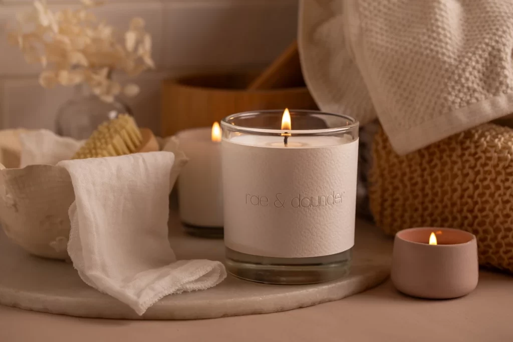 A lit rae & daunder candle on a marble board with soft cloths, a small tealight and natural bathroom accessories.